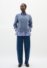 Svetlo modra pulover nad belo in modro črtasto srajco z nagubanimi robovi; v kombinaciji s temno modrimi širokimi hlačami in črnimi loaferji.