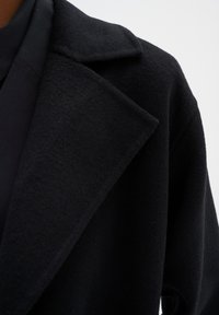 Manteau en laine noir avec un col cranté, texture douce et coupe ample. La vue rapprochée met en valeur les détails du tissu et le design du col.
