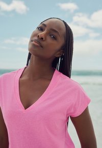 Roza majica s V-izrezom izrađena od mekanog, laganog tkanina s kratkim rukavima. Model ima ispletenu kosu i neutralnu pozadinu plavog neba i oceana.