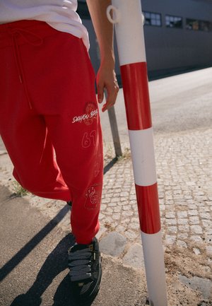 Rote Jogginghosen mit Kordelzugbund, die grafische Designs, einschließlich des Textes "DAY YOU BA" und eines Basketballmotivs, aufweisen, kombiniert mit schwarzen Sneakern.