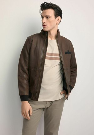 Brązowa skórzana kurtka bomberka z suwakiem z przodu, ściągaczami na mankietach i czarną naszywką. Noszona na jasnobeżowej koszulce z poziomymi paskami.