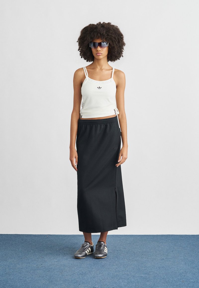 Débardeur blanc court avec liens réglables, jupe longue noire avec fermeture éclair sur le côté, chaussures de sport argentées à trois bandes, et lunettes de soleil foncées.