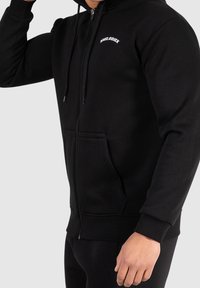 Felpa con zip nera realizzata in tessuto morbido. Presenta una tasca frontale e un logo sul petto. I polsini e l'orlo aderenti ne migliorano il design.