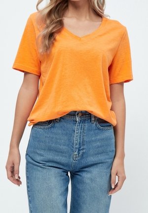 Femme portant un t-shirt orange vif à manches courtes et col en V, rentré dans un jean bleu taille haute sur un fond uni.