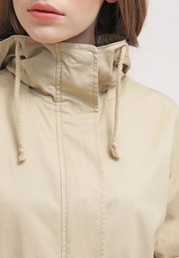 Beige jacka med hög krage och snörning, med en slät textur och minimalistisk design. Framdelen har dragkedjestängning utan synliga logotyper.