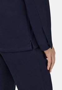 Mörkblå kostymjacka med slät textur, med en sidoventil och en synlig knapp på manschetten, i kombination med matchande byxor.