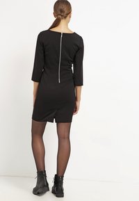 Black fitted dress with three-quarter sleeves, back zipper detail, above-knee length, and smooth texture. Paired with black lace-up ankle boots.