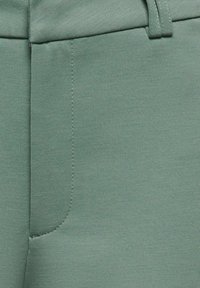 Tissu vert avec une texture lisse et une légère élasticité. Présente une poche passepoilée et des coutures le long de l'avant.