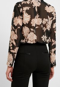 Przezroczysta czarna bluzka z beżowym kwiatowym wzorem, długimi rękawami i mankietami na guziki, w zestawieniu z czarnymi dopasowanymi spodniami z dyskretnym detalem kieszeni.