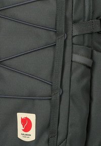 Tessuto dello zaino di colore grigio scuro con linee elastiche nere e una piccola etichetta bianca con il logo di una volpe rossa e il nome del marchio Fjällräven.