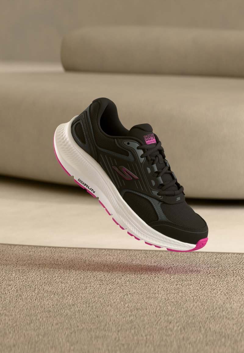 Chaussure de sport noire avec une tige en mesh, une semelle blanche et rose, et des accents texturés. Dotée de lacets et d'un logo sur le côté.