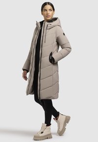 Beige gesteppter Puffer-Mantel mit Kapuze, mit seitlichen Taschen und schwarzem Besatz. Kombiniert mit cremefarbenen Ankle Boots und schwarzen Leggings.