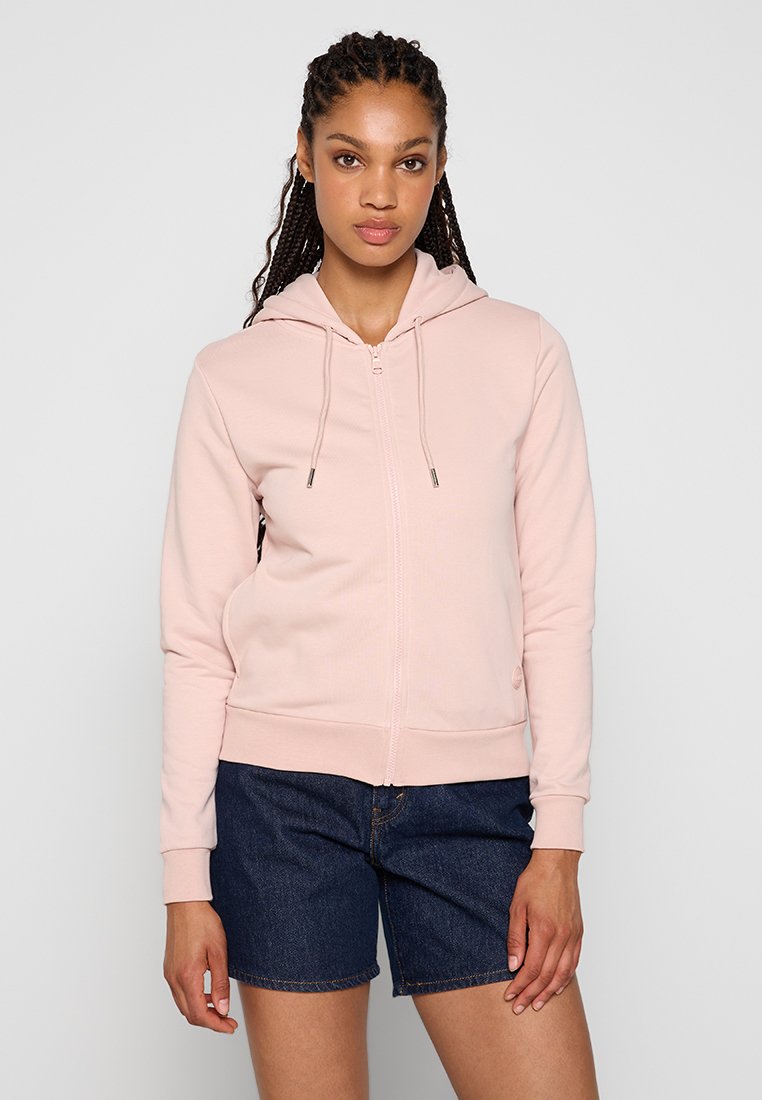 Felpa rosa chiaro con zip, dotata di cappuccio regolabile, polsini e orlo a coste, realizzata in tessuto morbido. Indossata sopra a pantaloni denim.