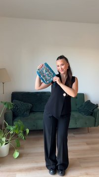 Turkis brodert clutch med blomsterdesign, rynkede kanter og håndledsstropp. Modellen har på seg svart antrekk mot en grønn sofa som bakgrunn.