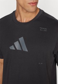 Camiseta de algodón negra con un logo triangular reflectante, el texto "TRAINING ALL_GYM" y un pequeño detalle "A_49" cerca del dobladillo.