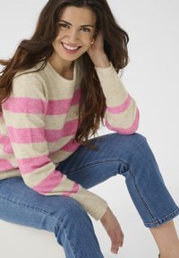 Beige sweater with pink stripes, round neckline, long sleeves, soft texture. Paired with blue cropped jeans, model seated against a light background.