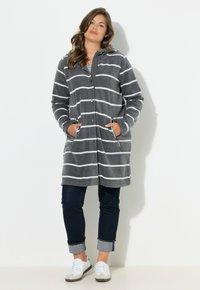 Cappotto grigio a righe, lungo fino a terra, con cappuccio, chiusura a bottoni, tasche laterali e texture morbida. Abbinalo a jeans scuri risvoltati e scarpe bianche.