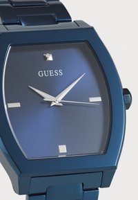 Montre en métal bleu avec un cadran carré, un cadran dégradé bleu, des aiguilles des heures et des minutes en argent, et un accent en diamant unique. Logo "GUESS" affiché.