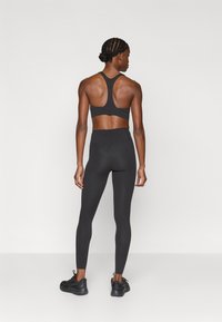 Atletyczna kobieta o splecionych włosach stoi tyłem, ubrana w czarny sportowy biustonosz, legginsy z wysokim stanem oraz czarne trampki na jednolitym tle.