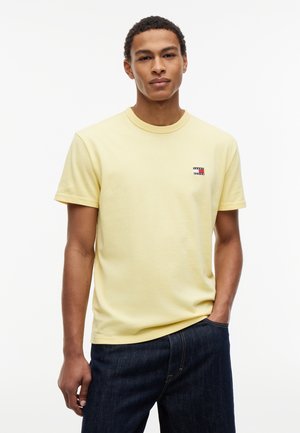 Junger Mann, der ein hellgelbes Kurzarm-T-Shirt mit kleinem Logo auf der Brust und dunkelblaue Jeans trägt, steht mit einer Hand in der Tasche.