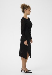 Crna čipkasta haljina s dugim rukavima, uskim siluetom i bočnim izrezom. Uparena s crnim potpeticama. Kovrčava kosa svezana u punđu.