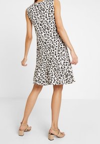 Ein ärmelloses, knielanges Kleid aus beigem Stoff mit einem schwarzen Leopardenmuster, mit einem rüschengesäumten Saum und lockerer Passform. Akzentuiert mit sandalen mit Riemen.