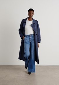Manteau trench bleu marine avec de grands revers, associé à un haut blanc ajusté et un jean bleu évasé. Le mannequin porte des escarpins noirs pointus.