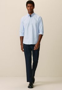 Himmelblaues Langarmhemd mit Button-Down-Kragen, kombiniert mit navyblauen Hosen. Der Stoff wirkt glatt mit einer subtilen Textur. Schwarze Schuhe runden das Outfit ab.
