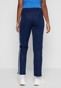 Tmavomodré polyesterové športové nohavice s bielymi bočnými pruhmi, zúženým strihom a elastickým pásom, doplnené bielymi športovými topánkami.