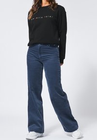 Mustat collegepaidat, joissa on teksti "AMERICA TODAY", yhdistettyinä sinisiin corduroy-levyyn housuihin. Valkoiset lenkkarit täydentävät asukokonaisuutta.