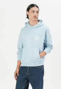 Lys blå hettegenser laget av myk stoff, med en frontlomme og hvit logo på brystet. Kombinert med vide denim-bukser.