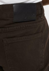 Pantalones de color marrón oscuro con una tela texturada, con un parche de cuero negro en la cintura con las letras "DEF" grabadas.