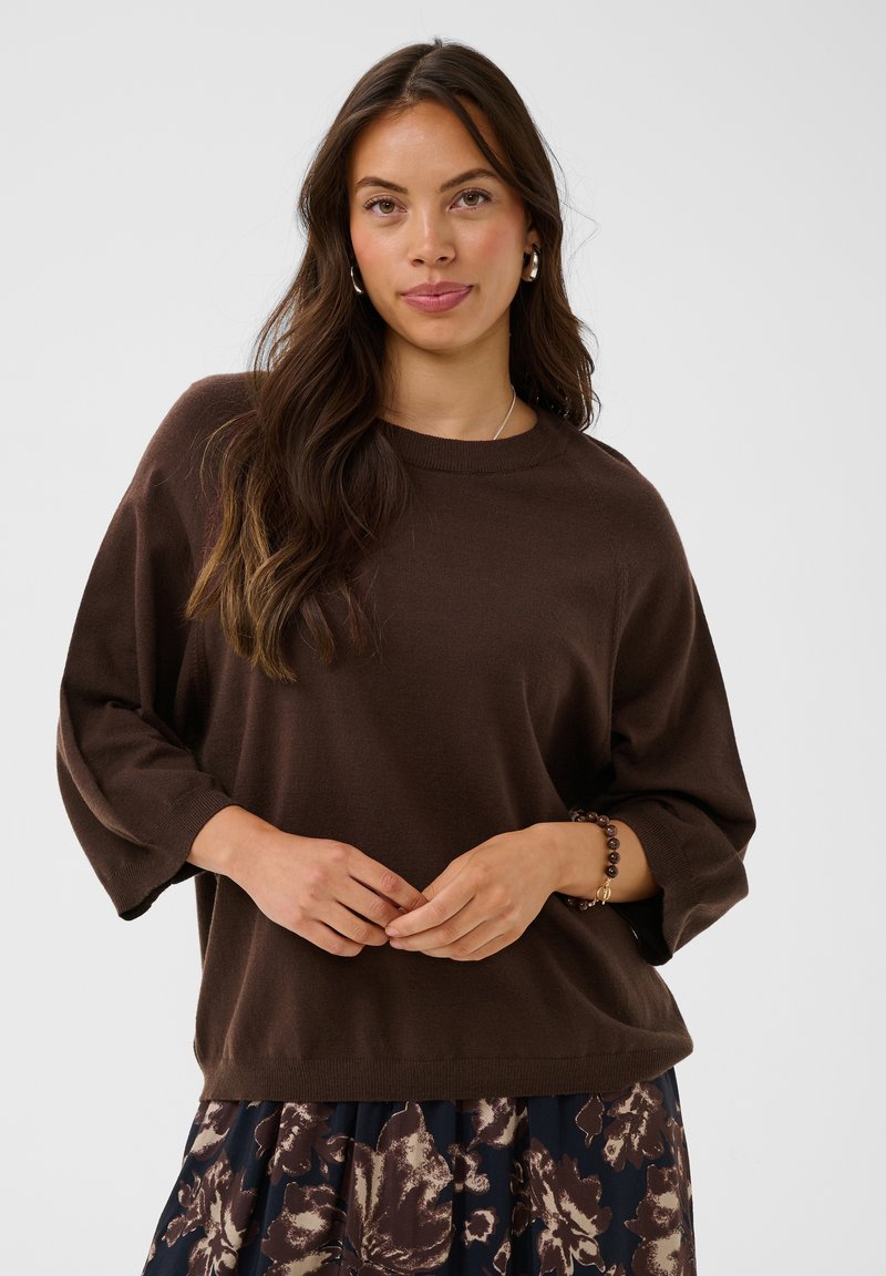 Jeune femme aux longs cheveux foncés portant un pull marron ample et une jupe fleurie, debout les mains jointes à la taille.