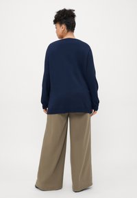 Γυναίκα με σγουρά μαλλιά που φοράει ναυτικό μπλε μακρυμάνικο πουλόβερ και ανοιχτόχρωμο παντελόνι φαρδιάς γραμμής, στέκεται με την πλάτη γυρισμένη σε μια απλή φόντο.