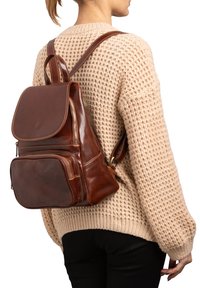 Zaino in pelle marrone con patta, numerose tasche con zip e cuciture rinforzate. Indossato sulla schiena sopra un maglione beige in maglia.