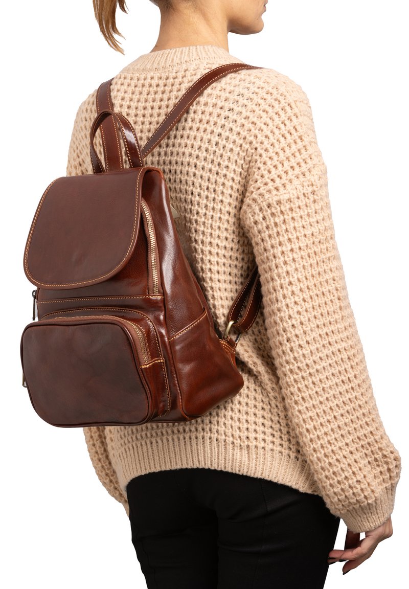 Zaino in pelle marrone con patta, numerose tasche con zip e cuciture rinforzate. Indossato sulla schiena sopra un maglione beige in maglia.