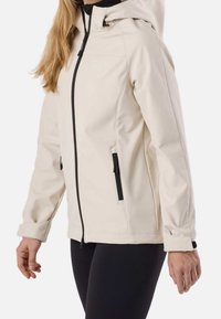 Femme portant une veste à capuche beige clair avec des fermetures éclair noires et un pantalon noir, debout devant un fond blanc.