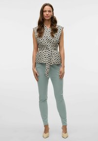 Ärmelloses Oberteil in Creme mit schwarzem Diamantenmuster, in der Taille gebunden. Kombiniert mit hellgrünen, eng anliegenden Hosen und beigen Blockabsatzschuhen.