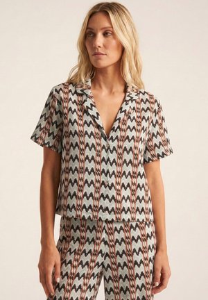 Mujer rubia con camisa de manga corta abotonada y pantalones a juego con rayas verticales en zigzag de color negro, blanco y marrón.