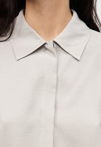 Blouse gris clair avec col classique, boutonnage sur le devant et texture douce. Coutures visibles le long des bords, éléments de design sans couture.