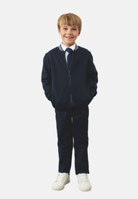 Marineblauwe bomberjack met rits en witte gestreepte kraag, wit shirt, marineblauwe das, bijpassende broek en witte sneakers. Casual uniformlook.