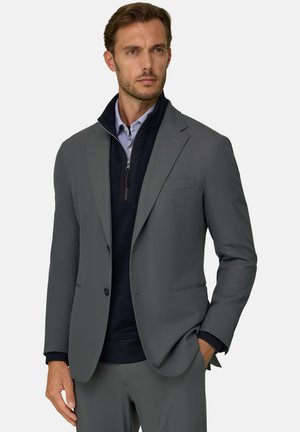 Hombre con una chaqueta de traje gris sobre un jersey azul marino con cremallera hasta un cuarto y una camisa de rayas azul claro, con una mano en el bolsillo, mirando hacia delante.