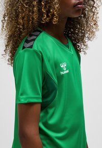 Grön sporttröja i slät tyg med V-ringning. Har svarta zickzackdetaljer på axlarna och en vit logga på framsidan.