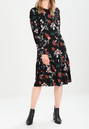 Frau in einem schwarzen Midikleid mit roten, weißen und grünen Blumenmustern, langen Ärmeln und schwarzen Stiefeletten, stehend vor einem weißen Hintergrund.
