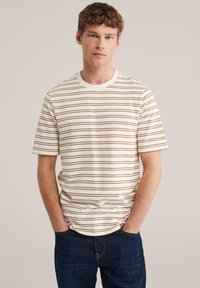 Ung mand med krøllet hår iført hvid t-shirt med tynde brune vandrette striber og mørkeblå jeans, hænderne i lommerne, neutral baggrund.