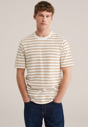 Jonge man met krullend haar, gekleed in een wit T-shirt met dunne bruine horizontale strepen en donkerblauwe spijkerbroek, handen in de zakken, neutrale achtergrond.