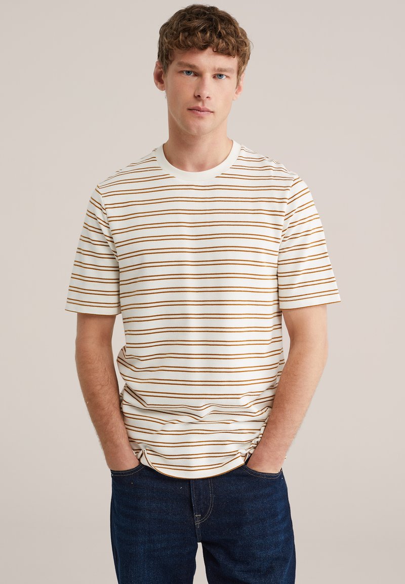 Ung mand med krøllet hår iført hvid t-shirt med tynde brune vandrette striber og mørkeblå jeans, hænderne i lommerne, neutral baggrund.