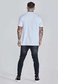 Bijela majica s okruglim izrezom, kratkih rukava, nošena uz crne traperice uskog kroja i crne tenisice. Glatki materijal, opušteni kroj, minimalni detalji.