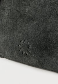 Κοντινή λήψη υφάσματος από μαύρο σουέντ με μικρό ανάγλυφο κυκλικό λογότυπο στο κέντρο κάτω μέρος σε λευκό φόντο.