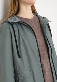 Femme portant une veste de pluie grise à capuche avec des cordons noirs par-dessus un pull en maille marron, regardant légèrement vers le haut et de côté, éloignée de la caméra.